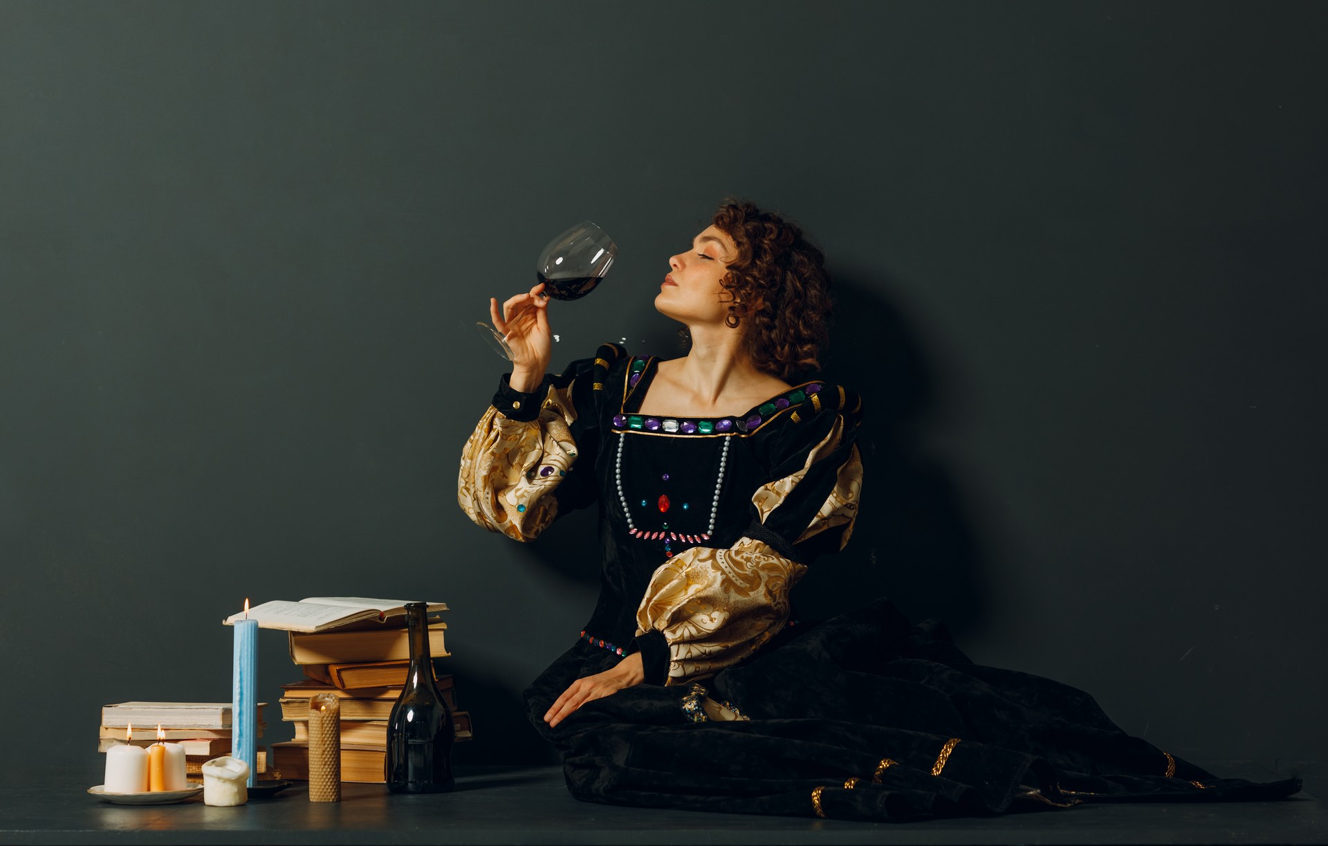 Young adult woman dressed in a medieval dress sitting near ancient books and candles with bottle of red wine and holding a glass of wine.